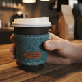 A reusable coffee cup with a white lid and a textured teal fabric sleeve, resting on a dark red textured surface. A small tan label on the sleeve reads 'Handmade'.